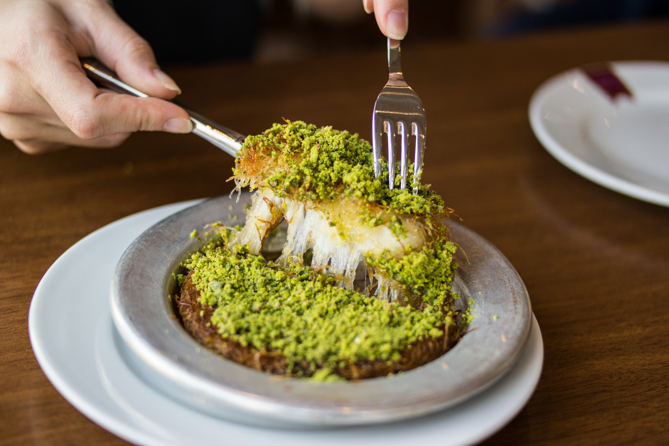 A warm, crispy dessert made with shredded phyllo dough, melted cheese, and sweet syrup, topped with pistachios